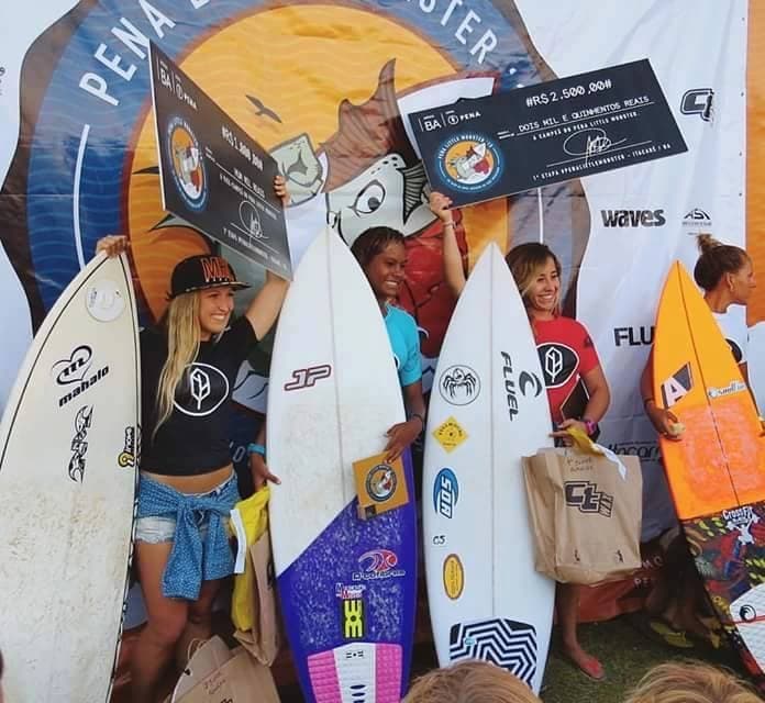 Marina Rezende in a red jersey on the podium of a women's surf competition, next to other athletes holding prize cheques.