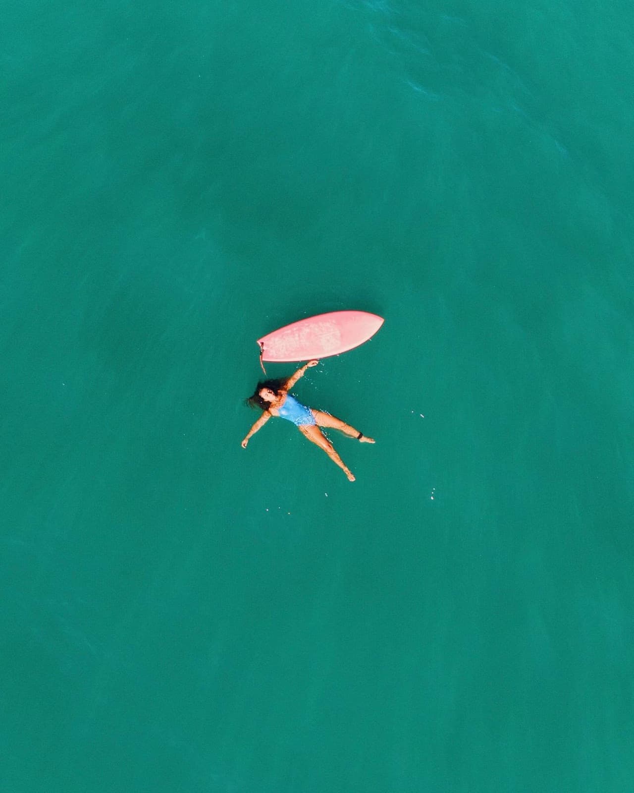 Aerial view of Marina Rezende floating on her back in blue-green water, next to a pink surfboard.