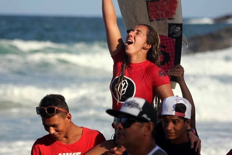 Marina Rezende celebrating a win on the beach, arm raised and holding her board, carried on someone's shoulders.