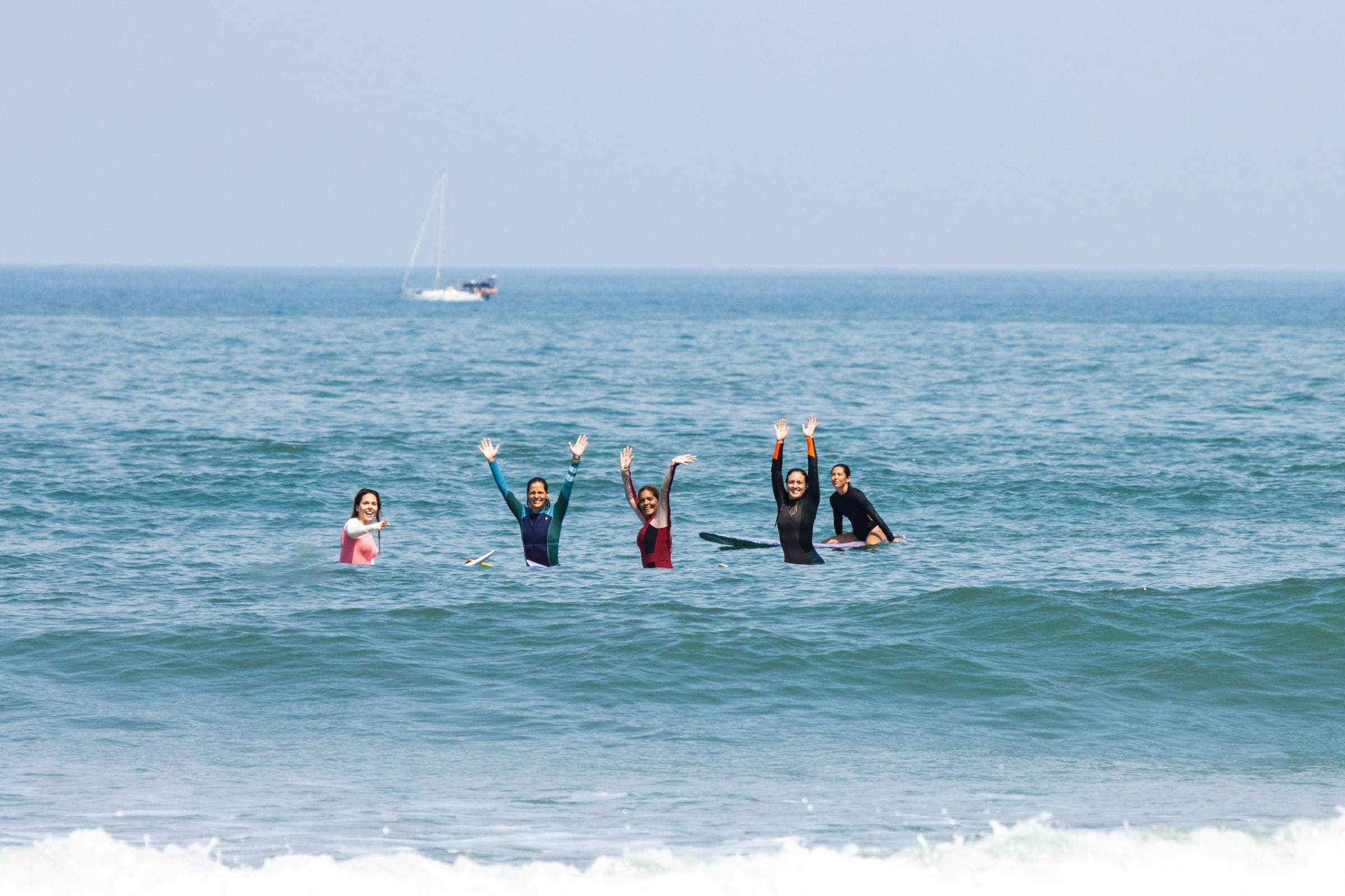 Five women in wetsuits waving from the ocean.