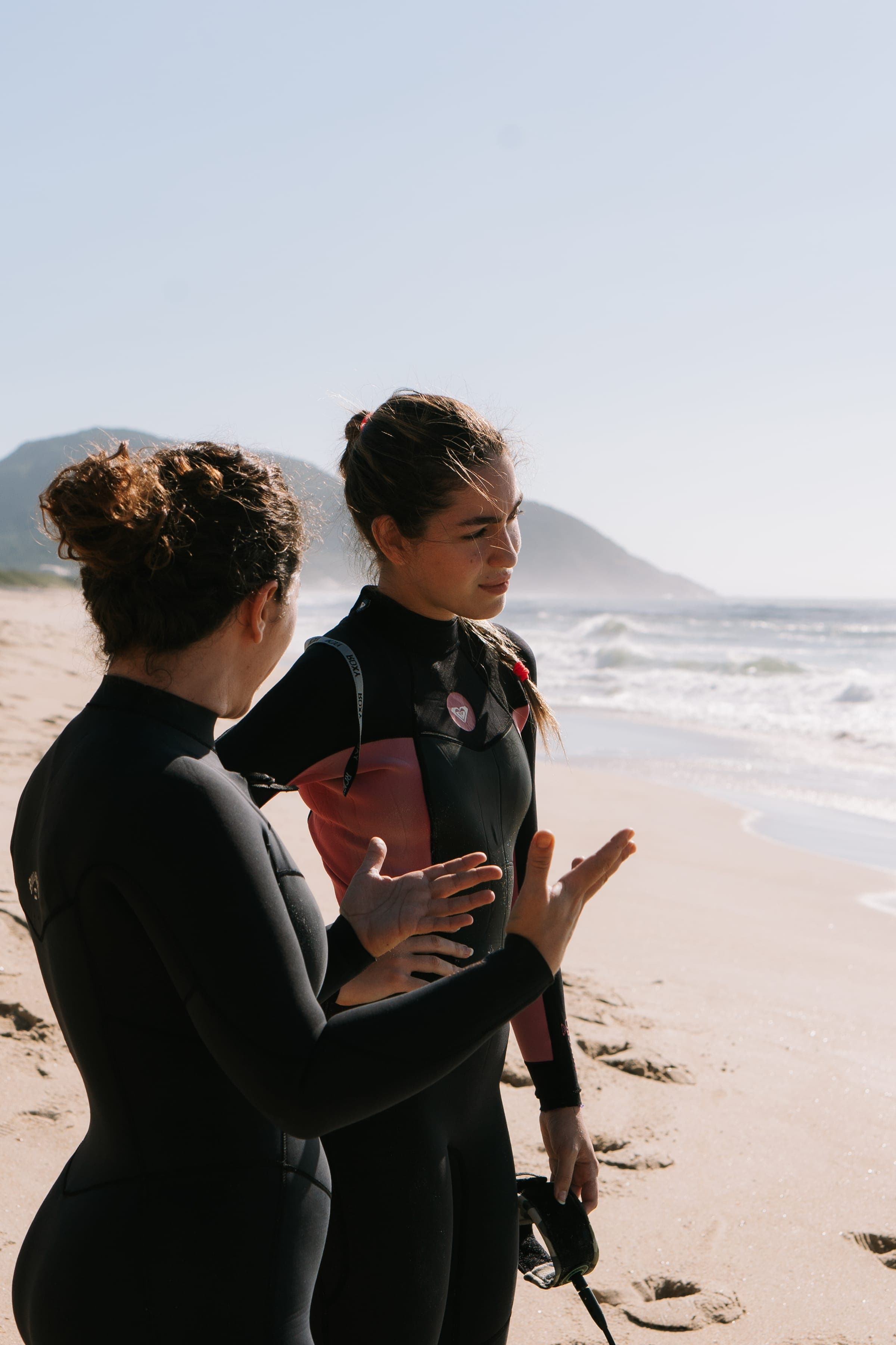 Two women in wetsuits talking on the beach, one gesturing as she explains something to the other.
