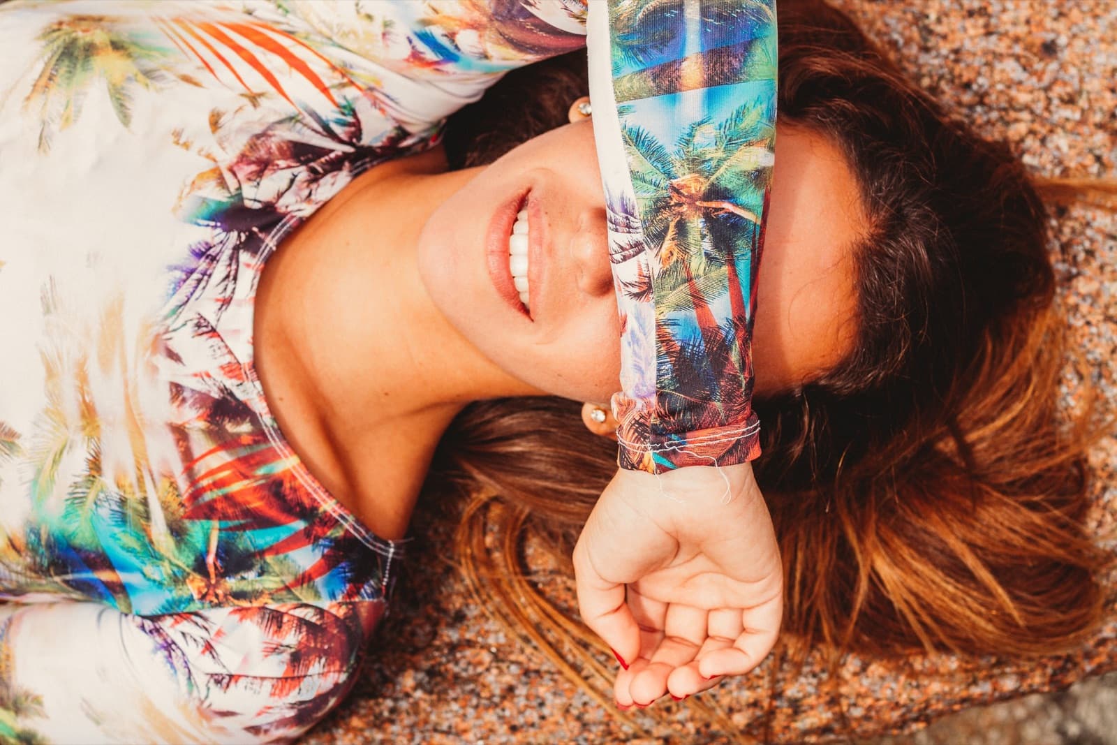 Marina Rezende lying on the sand, smiling, shading her eyes from the sun with her arm and a palm-print shirt.