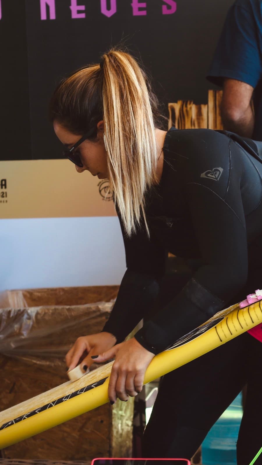 A surfer in a wetsuit and sunglasses, with a sun-bleached blonde ponytail, waxing a yellow surfboard.