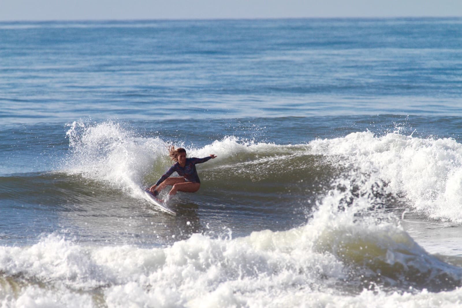 A surfer in a long-sleeved rash guard carving on a wave through the spray, on a sunny day with a blue sea.