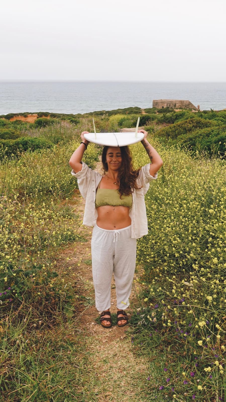 Marina Rezende walking along a path between yellow flowers, a white surfboard balanced on her head and the sea behind her.