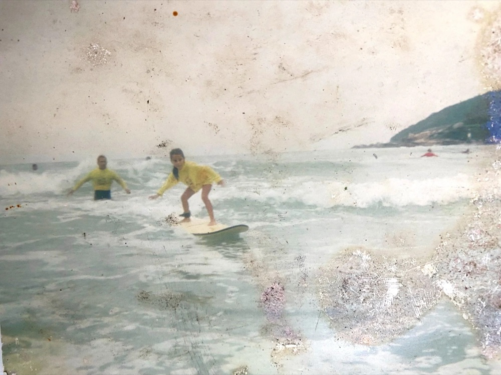 Old photo of a young girl surfing a small wave, with an instructor beside her