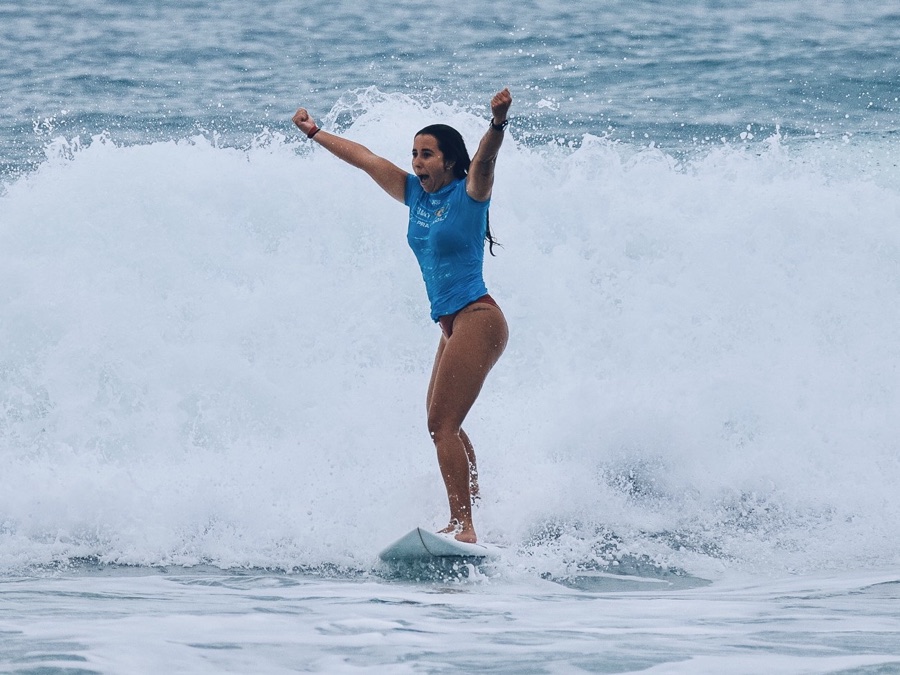 A surfer in a blue rash guard riding a wave with both arms raised